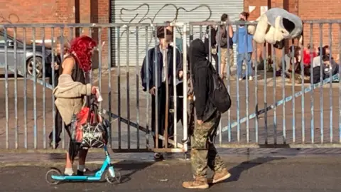 About a dozen young people walking in daylight outside the former Wickes building, with graffiti on a large garage entrance in the background and steel gates in the foreground