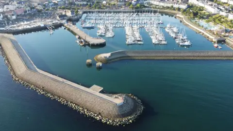 An aerial view of a large number of boats moored in Bangor's marina. It is a bright sunny day and the Eisenhower Pier is in the foreground. The city of Bangor can be seen on the edges of the picture which is dominated by sea.