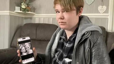 BBC A woman on a sofa is looking at the camera and holding a mobile phone, which has a photo on it.