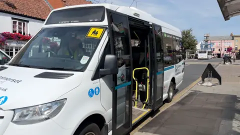 A minibus parked at a bus stop in a town centre. In the distance there are colourful buildings.