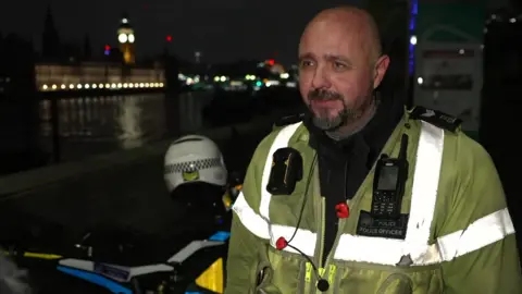 Sgt Ryan Perry in a high visibility jacket stands next to one of the bikes, his police helmet resting on top of it. Behind him is the River Thames and Houses of Parliament 