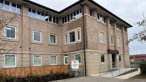 The entrance to Cumbria coroner's court building. The entrance is at the left-hand side of a three-storey building, with beige cladding on the ground floor and shelldash on the upper floors. There is a sign for the coroner's court in front on a ramp leading to the door.