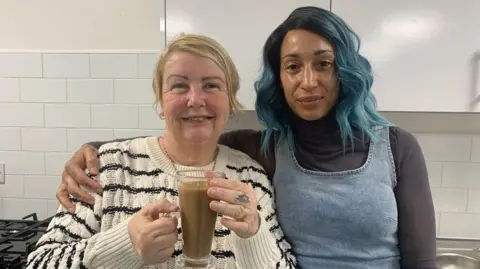 Two women in a kitchen stand shoulder to shoulder. Diane Wood is holding a glass coffee cup, the other, volunteer Rachel Wilson, has her arm around her.