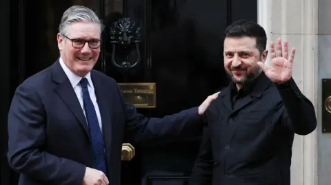 President Volodymyr Zelensky waves at cameras outside the black door of 10 Downing Street as he is welcomed by Prime Minister Sir Keir Starmer, who places his hand on his shoulder.