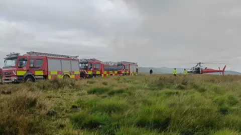 NWFRS Fire engines and a firefighting helicopter in a field with mountains in the background. The helicopter is white in the front and red in the back. 
