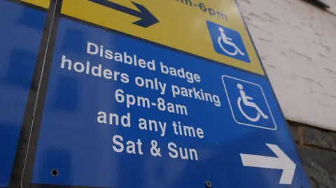 BBC A disabled badge holder sign. The bottom half is blue and has the words 'Disabled badge holders only parking 6pm-8am and any time Sat & Sun'. There is a disabled badge logo to the left and an arrow pointing to the right. Above is part of a yellow sign with the same logo.