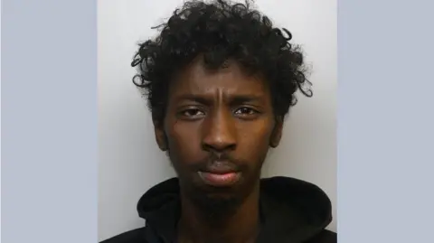 Mugshot of Abdinasir Hussein. He is a 24-year-old Black man with short-medium curly black hair, and is wearing a black hoodie.