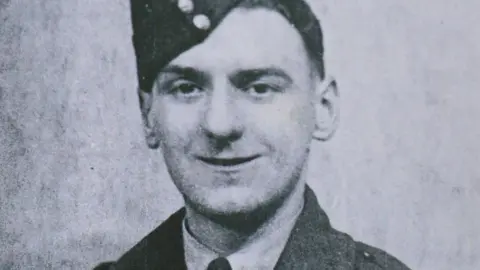 Bernard Morgan A black and white photo of a man in military uniform. He has a soft cap perched slightly on the right side of his head. He is clean shaven with dark hair and the top of a dark jacket can be seen over the white collar on a shirt.