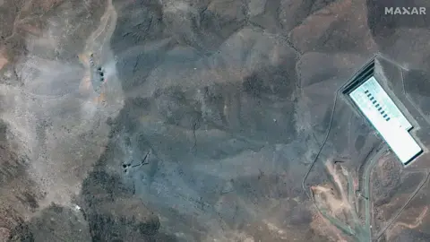 Maxar Satellite Imagery shows the ridge above the Fordo complex post-strike. Multiple impact craters and a wide grey-blue ash layer are visible across the ridge, suggesting direct hits to the overburden covering the underground facility. 