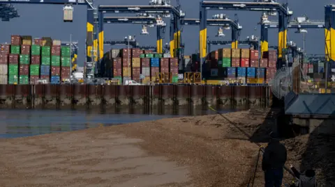 Reuters Port of Felixstowe wide showing cranes and freight with a sand bank in the foreground and a fisherman watching on