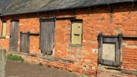 Google An image of the horse stables. It is an old red brick building with wooden doors and shutters, some of which are worn at the edges.
