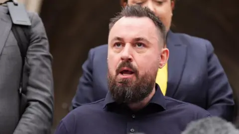 Martin Hibbert sits speaking. A media microphone is in front of him and people stand behind him. 