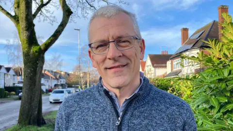 An elderly man with short white hair and thin framed glasses. He is wearing a blue fleece and is stood on a residential street.