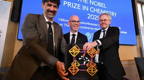 Getty Images Three members of the Nobel committee hold a model of metal-organic frameworks, which is an orange, pink and yellow structure.