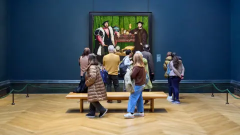 David Levene/Art Fund/PA Media People stand in front and behind a bench looking at a giant Tudor-style painting of two men. The gallery has a dark blue wall.