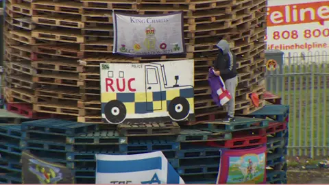 A youth climbs onto a bonfire to place a flag on it, a number of flags including the Israel flag are already on it