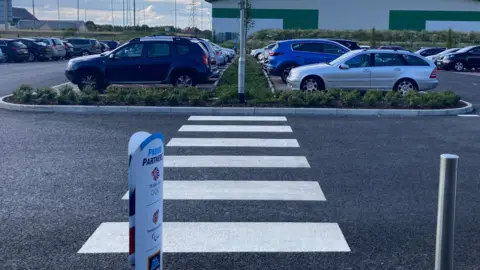 A car park outside an Aldi store. The zebra crossing, white of black tarmac, leads to a kerbed flower bed instead of a footpath. The car park is almost full. There is a warehouse-type building in the background. The sky is cloudy.