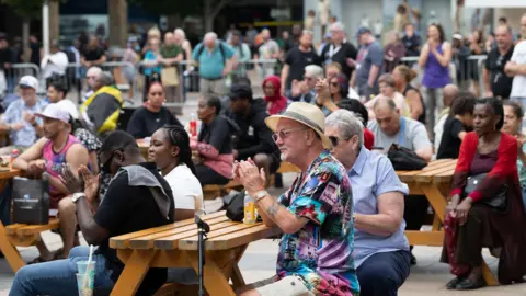 Crowds of people clapping during a performance. They are sitting on benches infront of a main stage