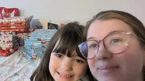 Kirsty Barker Kirsty and her daughter in a selfie image with a pile of wrapped Christmas presents behind them. Kirsty is wearing glasses and has a nose piercing. Her brown hair is tied back while her daughter's dark hair is down and she has a fringe.
