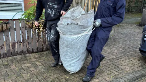 A man in a navy boiler suit, wearing black boots, hold a large white sack with both his hands, which are protected by grey gloves. He stands to the right of the bag. To the left of the bag is a man in a black hoodie, black trousers and boots. They walk in front of a dark-grey fence, on a grey brick driveway. 