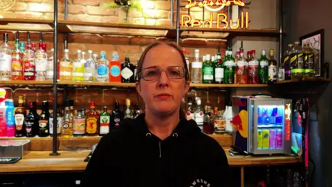 Chris McHugh/BBC Maria Kerry from The Globe Inn posing for an image. The bar with a multitude of bottles is behind her. There is also a small fridge filled with energy drinks. She is wearing glasses.
