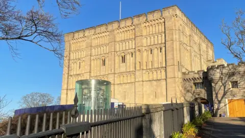 Shaun Whitmore/BBC An external view of Norwich Castle, which was built by sandstone. A glass elevator is located in front of the castle and there is a bright blue sky.