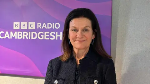 Sue Dougan/BBC Bridget Smith stares at the camera. She is wearing a black jacket and has shoulder length dark hair. She stands in front of a sign that says BBC Radio Cambridgeshire. 