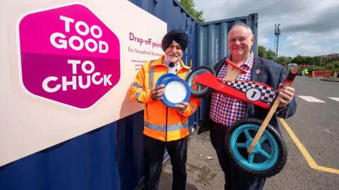 Two men, one holding plates and another holding a small, wooden kids bike, standing next to a blue shipping container at the side of a road. A pink posted is on the side of the container and says "Too Good To Chuck". 