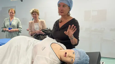 Eluned Morgan stood partially in scrubs in front of a model of a pregnant woman, lying on a bed