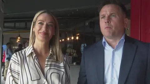 Jackie Read has long blond hair and is wearing a white and black blouse.
Brian Reid has short dark hair and is wearing a dark grey suit jacket and light blue shirt