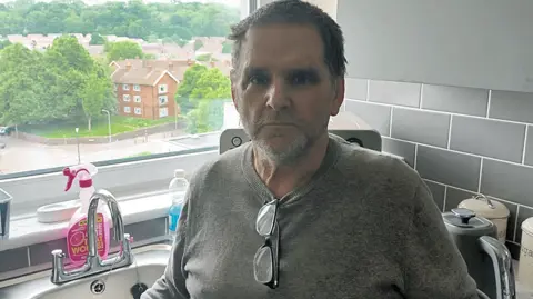Martin Newman next to the sink in his flat with a window behind him at Caerau Court, Cardiff. He has short hair and short grey stubble and is wearing a grey jumper. 