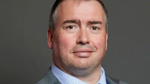 UK Parliament A man with grey hair, wearing a blue and white striped collared shirt and a grey blazer and looking at the camera with a half-smile.