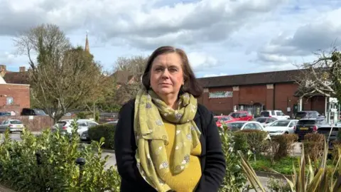 A woman - Councillor Elizabeth Lee - standing with her hands together in front of her wearing a black cardigan and an olive-coloured scarf, standing next to a car park with cars and brick buildings in the background. 