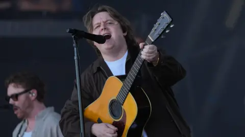 PA Media Lewis Capaldi is on stage with an accoustic guitar singing into a microphone. He has long hair and he is wearing a black jacket.