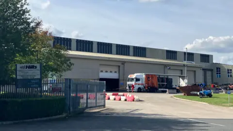 A lorry outside a building with a grey fence and a sign which says 'Hills'
