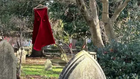 Ros Martin A red velvet dress hanging on a hanger from a tree in a churchyard with the tip of a large gravestone in the foreground