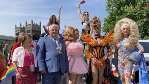 STUART PRANDLE/BBC Mayor Gary Hutchinson, wearing a blue suit and his golden chain of office, stood with three drag queens taking part in the march. They wear dramatic dresses. Notably, one wears a dress designed to look like orange flames, with a golden crown fashioned to look like a blooming flower.