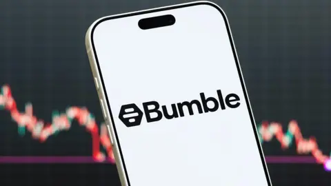 A close-up of a white smartphone screen displaying the black Bumble logo, held against a blurred background showing a financial stock chart with red and green lines.