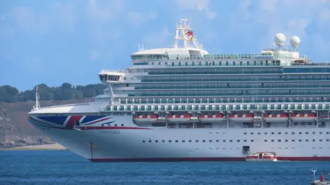 BBC This is a photo of a large cruise ship in the sea in front of some land. The cruise ship is white with the Union Jack flag on the front of the vessel. It bears the name 'Ventura'. 