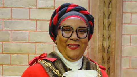 A woman wearing a red civil mayoral gown, white lace collar, glasses, hoop earrings and a red and black headdress smiles at the camera.