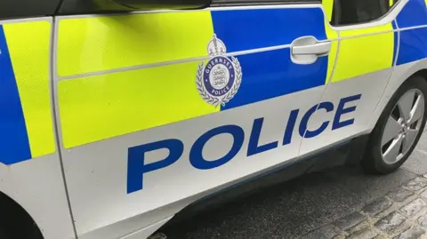 BBC Guernsey Police car pictured. The image only shows the door which is coloured yellow, blue and white with police in blue lettering. There is also a Guernsey Police emblem on the door.