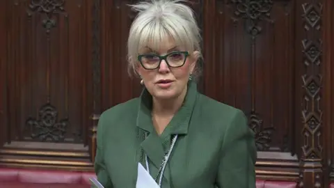 PA Media A womanin a green jackett and green glasses, speaks inthe House of Lords. She has white blonde hair which is tied up in a bun.