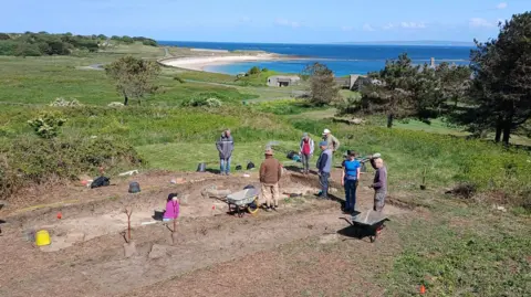 Dig Alderney A number of people at an archaeology excavation with a beach in the background.
