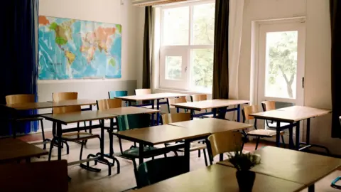 Photo of empty classroom with map of the world on the wall at the back. The classroom is tidy and all the chairs are tucked into the tables. There are large windows with trees showing on the far side. 