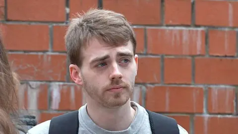 Spindrift Kyle Waddell has short, brown hair and is wearing a grey sweatshirt with a rucksack over his shoulders. There is a red brick wall behind him.