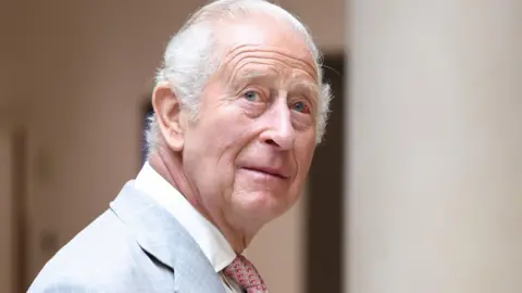 King Charles III looks up while wearing a grey suit, white shirt and red patterned tie