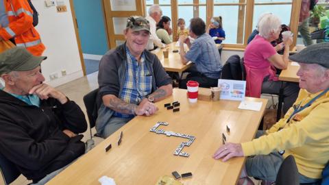 Three older men sitting round a table playing dominoes and smiling, with more tables in the background with more people sitting round them
