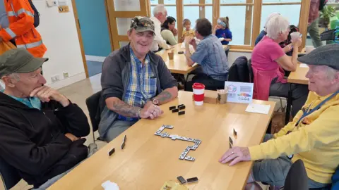 Educafe Three older men sitting round a table playing dominoes and smiling, with more tables in the background with more people sitting round them