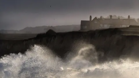 Getty Images Large waves crashing on sandy cliffs with cliff-top homes in the background