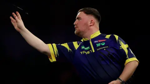 Getty Images Luke Littler throwing a dart. He has short brown hair and light beard. He is wearing a purple and yellow top. His non-throwing hand is down by his side.